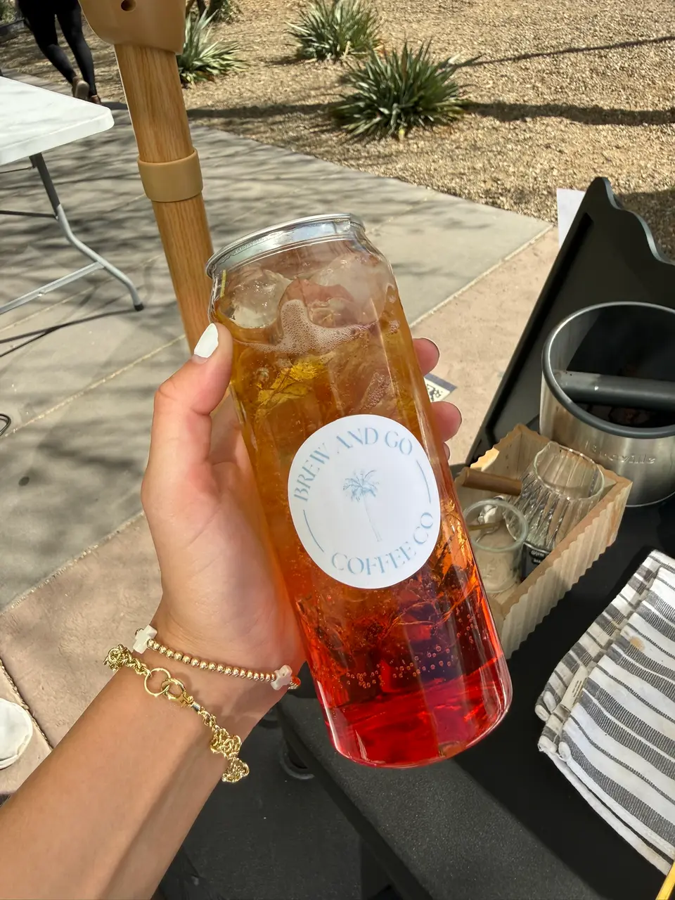 An amber and red iced drink in a Brew and Go can, held up beside her bar outdoors
