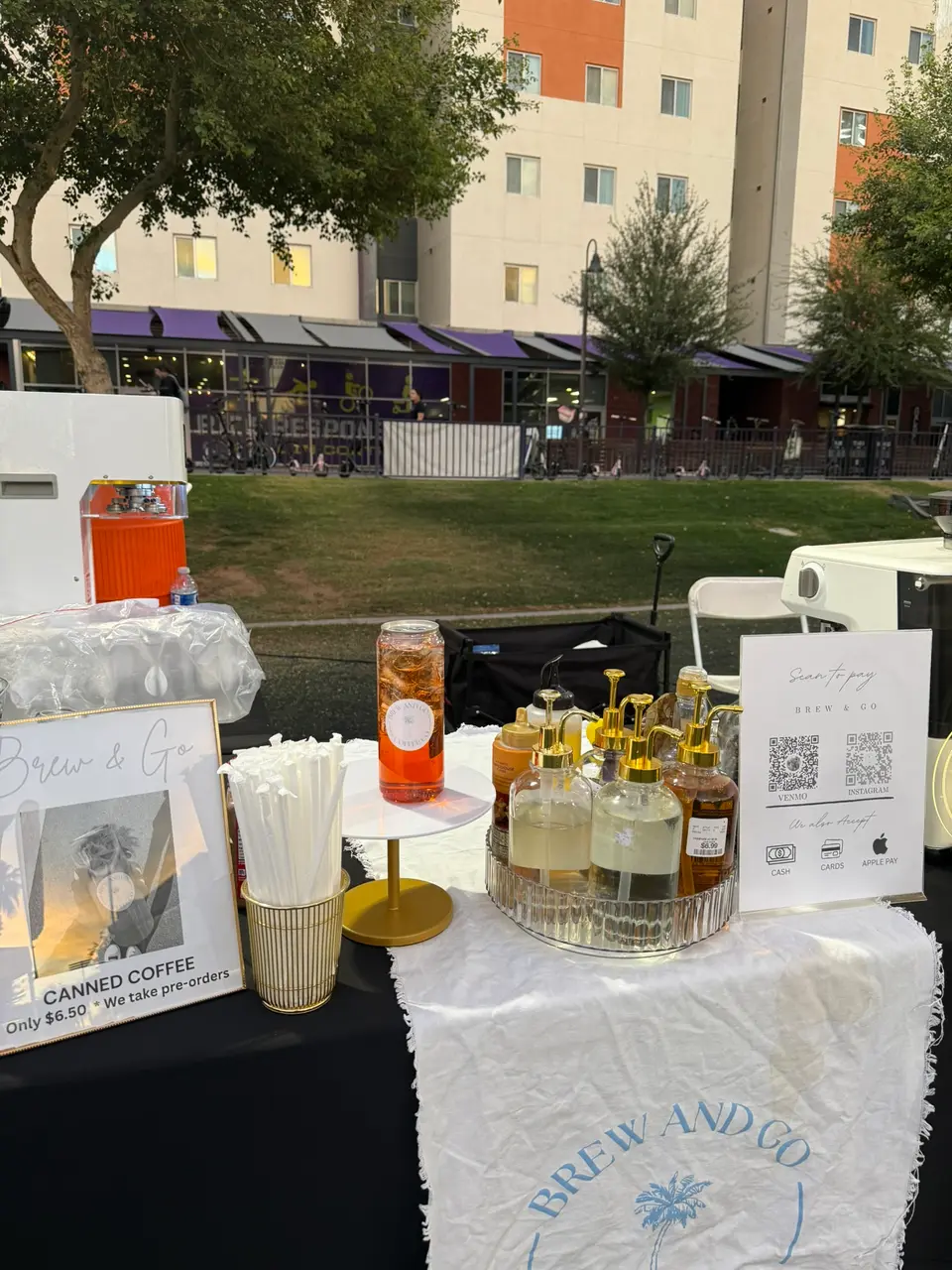 Her coffee bar on a lawn: syrups, straws, a canned-coffee sign and the Brew and Go cloth